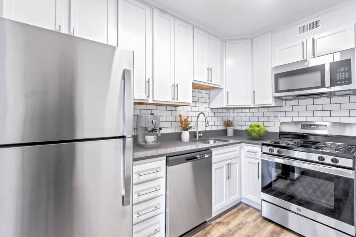 Modern kitchen with stainless steel appliances, white cabinets, subway tile backsplash, and wood flooring—perfect for those exploring apartment floor plans in Chevy Chase. A bowl of green apples adds a fresh touch on the counter.